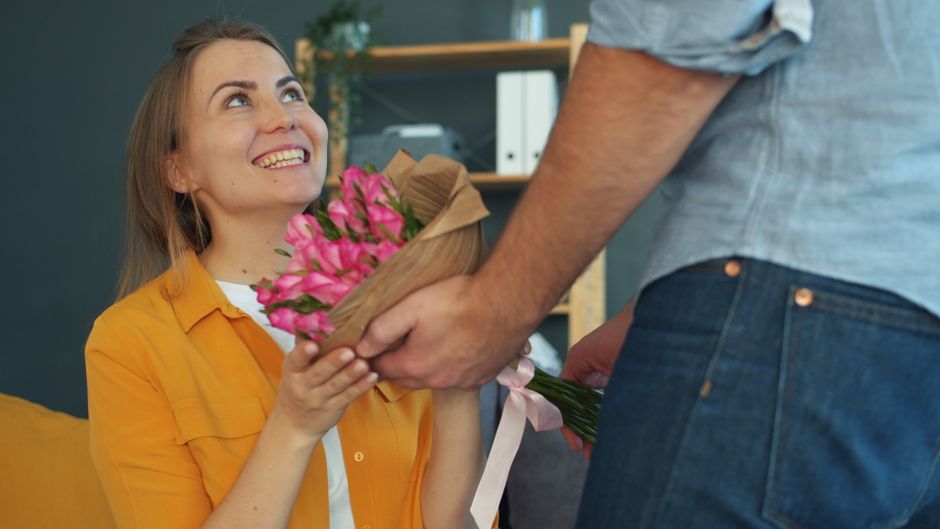 A young woman with long, light brown hair, smiling warmly and wearing a yellow shirt, is accepting a bouquet of fresh pink flowers wrapped in brown paper from a person whose torso and arm are visible,
