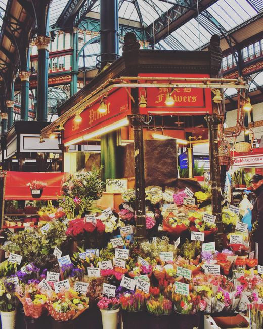 A vibrant floral display at a busy indoor market showcases an array of freshly cut flowers arranged in various bouquets and wrapped in colorful paper. The selection includes red, pink, purple, and yel