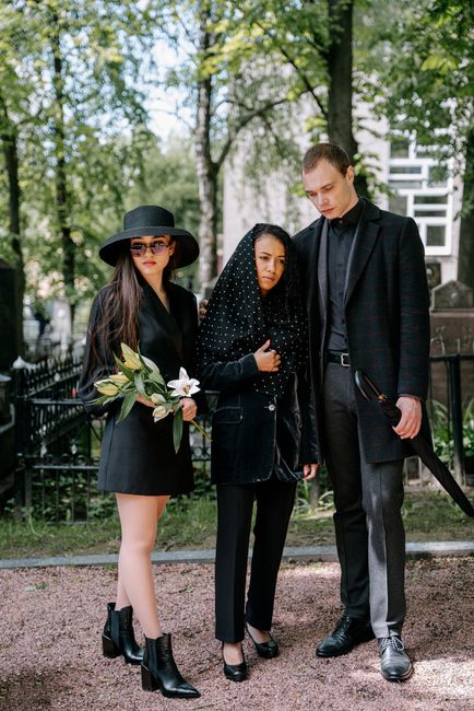 A group of three people standing outdoors in a park with lush green trees and a black metal fence in the background. The woman on the left is holding a bouquet of fresh flowers, including large white 