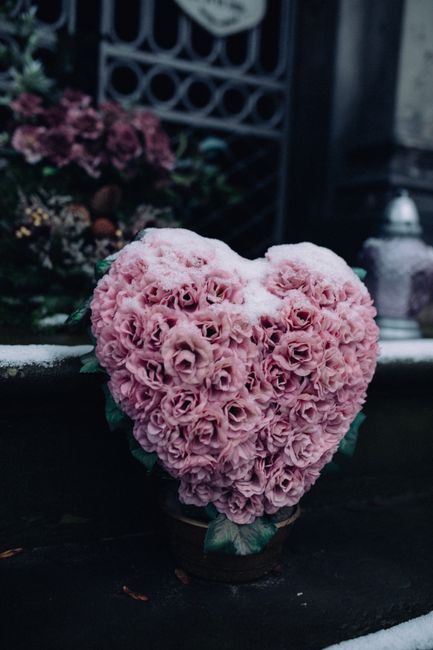 A heart-shaped floral arrangement composed of tightly clustered pink roses, creating a soft and textured appearance. The bouquet is placed in a small, dark-colored pot and is set on a dark surface, wi
