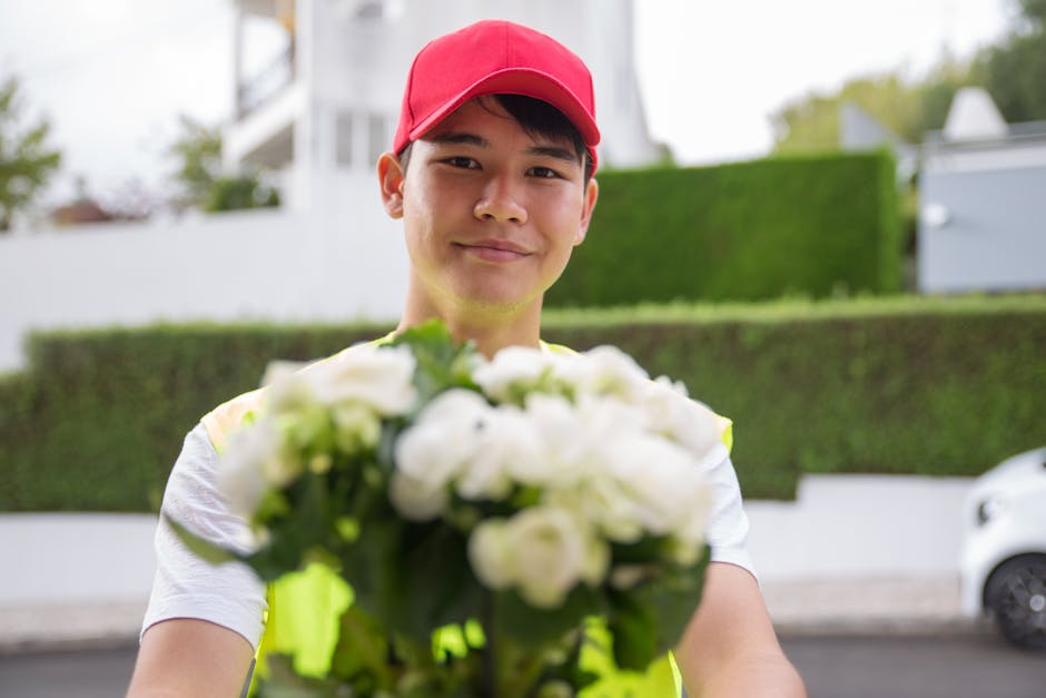 A delivery person wearing a bright yellow vest and a red cap is holding out a bouquet of fresh white flowers towards the camera. The bouquet features a mix of white roses, peonies, and ranunculus, ele