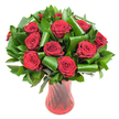 A lush, rounded bouquet of a dozen or more deep crimson roses fills the frame, each bloom at the peak of its velvet-soft perfection. The petals are tightly spiralled at the centre and gradually unfurl into rich, wine-dark cups, their velvety texture catching soft, even light that reveals subtle ridges and delicate edges. Between the rose heads a generous sweep of glossy dark green foliage creates a dense, verdant backdrop: long ruscus- or salal-like leaves form a thick collar, while a few broader, Aspidistra-style leaves have been skillfully rolled and curved into polished, architectural loops that cradle and guide the eye around the composition. Those sculpted leaves lend a contemporary, almost sculptural quality against the classic romance of the roses. The stems sit in a tall, translucent ruby-red glass vase with a faintly twisted or ribbed surface; vertical lines in the glass catch highlights and deepen the red tones so the vase seems to glow from within. Everything is set against a pristine white background, the simple backdrop making the rich reds and deep greens read with dramatic clarity and focusing attention on craft and texture. There's an immediacy to the image-a subtle scent of classic rose perfume seems to hover, and the arrangement reads equally as an intimate anniversary gift or an elegant centerpiece for a dinner after a walk on Blackheath Village. The composition feels timeless and composed, a balance of passionate colour and considered structure that a local florist might recommend for a heartfelt declaration or a quietly luxurious celebration in the Blackheath neighbourhood.