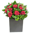 In this elegantly modern composition, rich crimson roses take centre stage, each bloom unfolding in velvety spirals that catch the light with a soft, satin sheen. The roses are closely clustered, their full heads rising like small red suns above an artful basket of glossy green foliage; each leaf has been gently curled and tucked to create a rhythmic pattern of movement and depth that both supports and accentuates the flowers. The arrangement sits in a crisp matte black cube, its geometric silhouette grounding the composition and lending a contemporary, gallery-like presence to the display. Lighting is clear and steady, picking out highlights on the petals and the polished surfaces of the leaves, and suggesting the cool clarity of a bright London morning or a clean, modern interior in Blackheath Village. Close to the blooms you sense a delicate, classic rose scent tempered by the green, almost citrus-bright notes of fresh foliage - a fragrance that feels clean and uplifting rather than heavy. Texturally, the contrast is tactile: the petals are plush and yielding while the leaves are firm and slightly waxed, folded into near-sculptural shapes by an experienced hand. This design reads as a statement of refined passion and calm order, appropriate for anniversaries, a heartfelt declaration, or to bring composed drama to a contemporary home or office near Greenwich Park. As a local florist would tell you, it's the kind of piece that looks equally at home on a Blackheath dining table or as a thoughtful same-day delivery for someone walking past the heath on a crisp afternoon.
